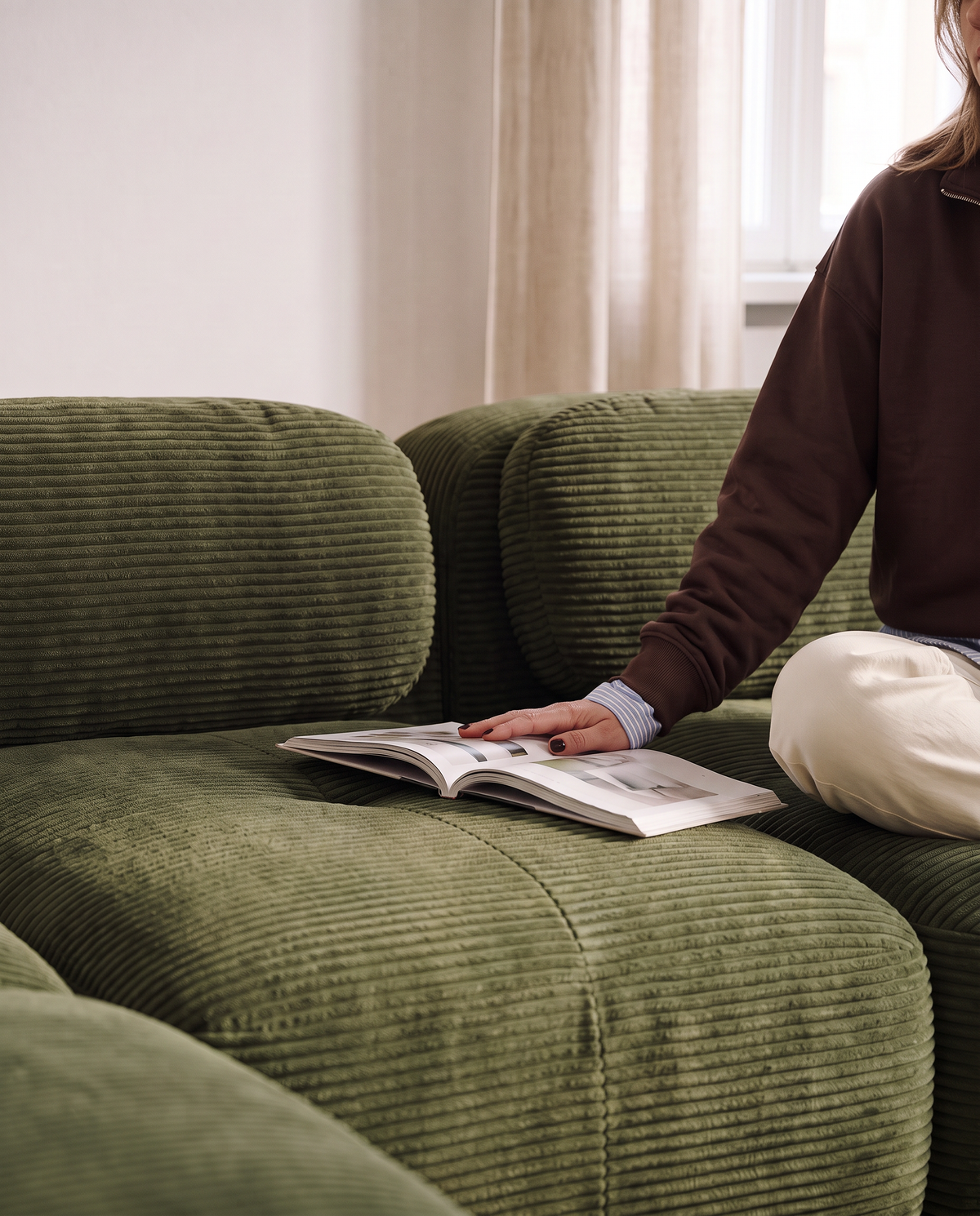 Femme assise sur le canapé Archipel en velours côtelé vert, un magazine à la main
